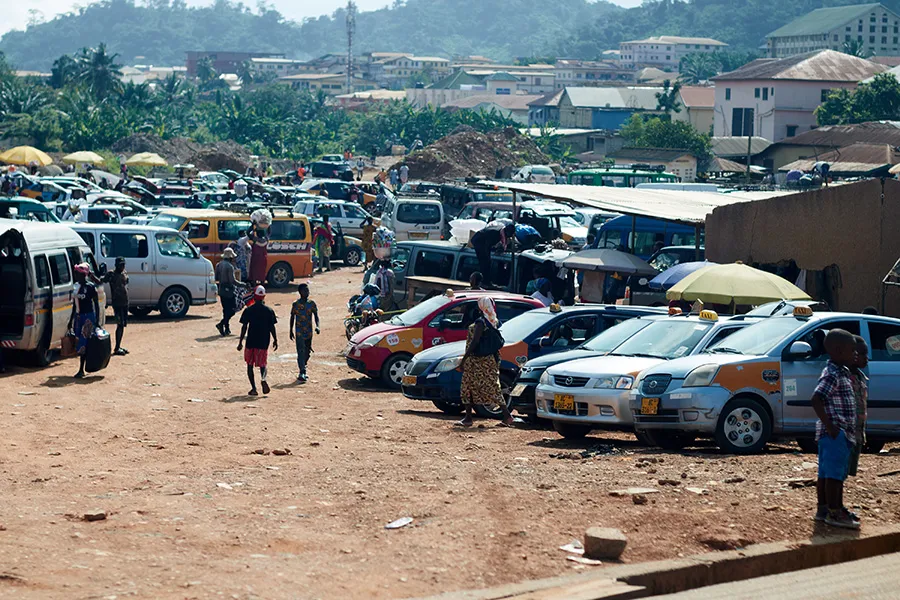 Image shows a road in Ghana with parked cars on the edge of the village